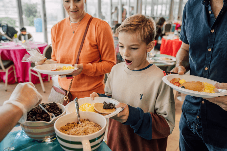 A family enjoys a buffet breakfast with a variety of food options, including pancakes, scrambled eggs, and toppings like nuts and fruit