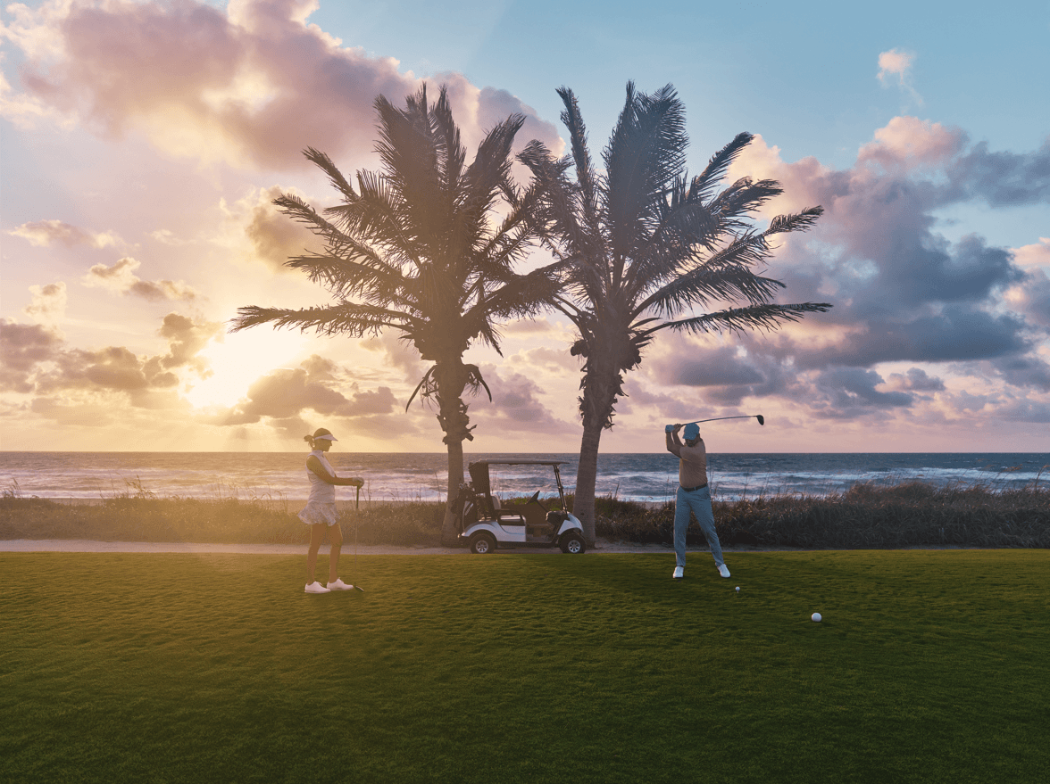 A golfer and caddy enjoy a serene sunset golf session on a lush green course by the ocean, framed by palm trees