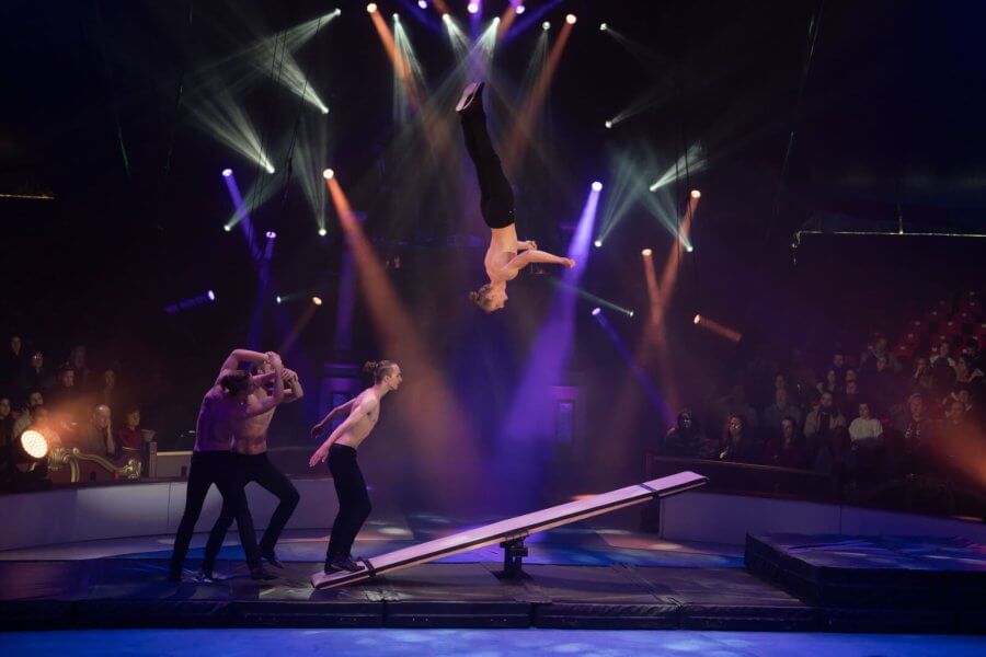 A circus performer executes a daring mid-air flip on a tightrope while three others assist below, all under a dazzling display of stage lights
