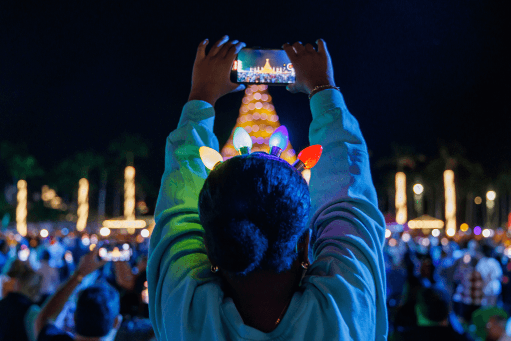 A person with colorful Christmas lights in their hair takes a selfie in front of a large, illuminated Christmas tree surrounded by a crowd at night