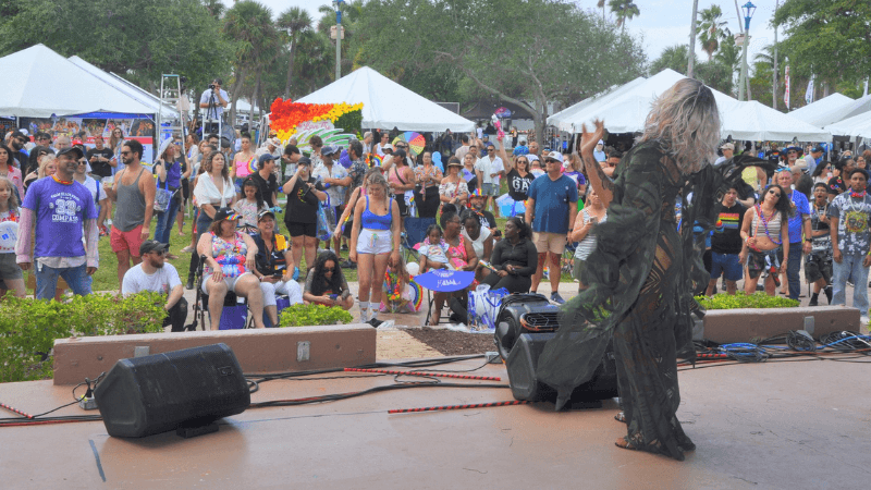 A performer in a flowing black dress dances energetically on stage at a vibrant outdoor festival, surrounded by a diverse crowd under colorful tents