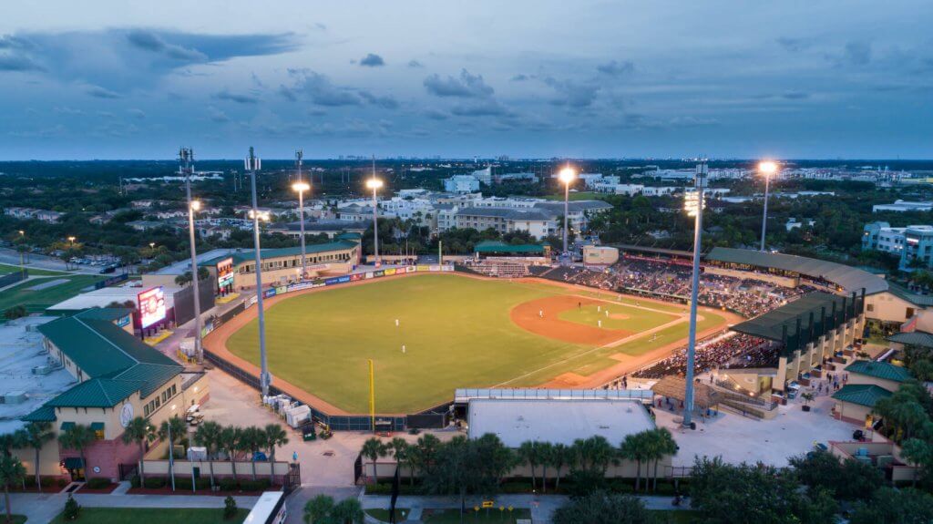 Aerial view of a baseball stadium at dusk, with the field illuminated and players on the diamond