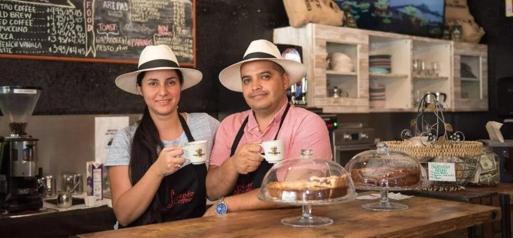 Deux baristas portant des chapeaux et des tabliers blancs se tiennent derrière un comptoir de café, chacun tenant une tasse de café et souriant