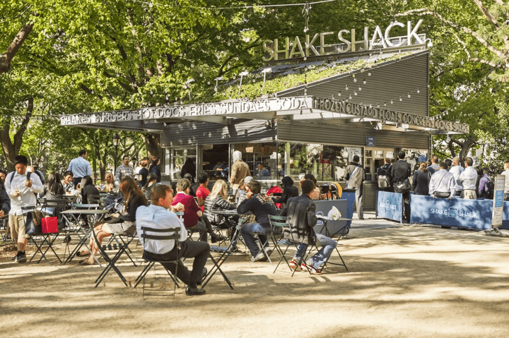 People enjoying burgers and shakes at an outdoor Shake Shack under a tree-covered park