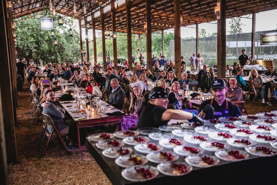 A lively outdoor dinner party with guests seated at long wooden tables under a rustic wooden pavilion, enjoying a meal with candles and flowers