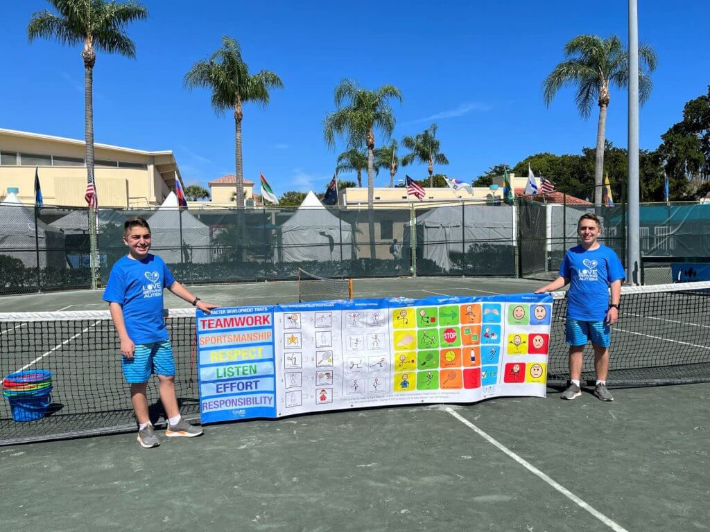 Two young boys stand on a tennis court holding a colorful banner promoting teamwork and sportsmanship