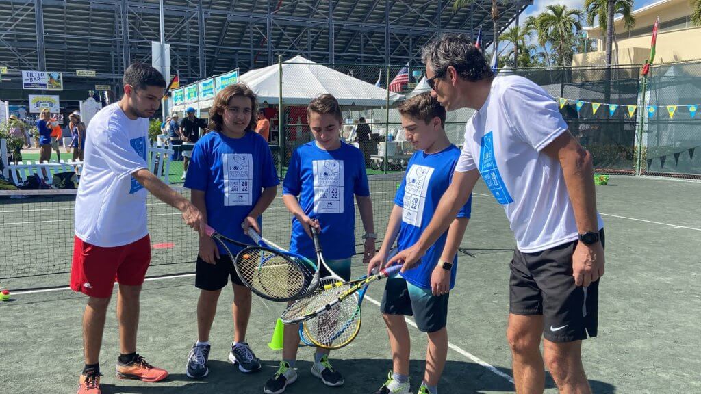 A group of five individuals, including four young boys and one adult man, are gathered on a tennis court, each holding a tennis racket and engaging in a tennis lesson
