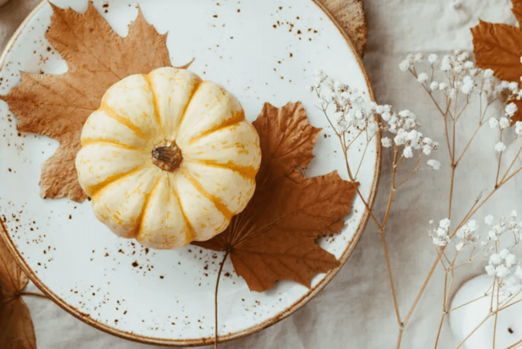 A small yellow pumpkin with white stripes sits on a white plate adorned with autumn leaves and delicate white flowers