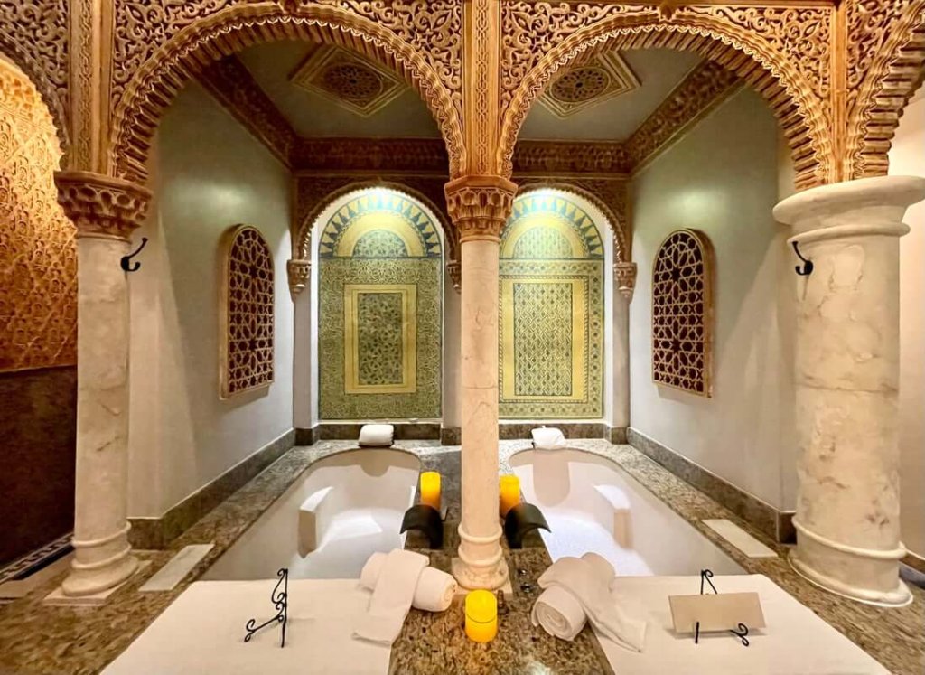 An opulent bathroom featuring twin marble bathtubs under intricately carved arches, adorned with candles and white towels