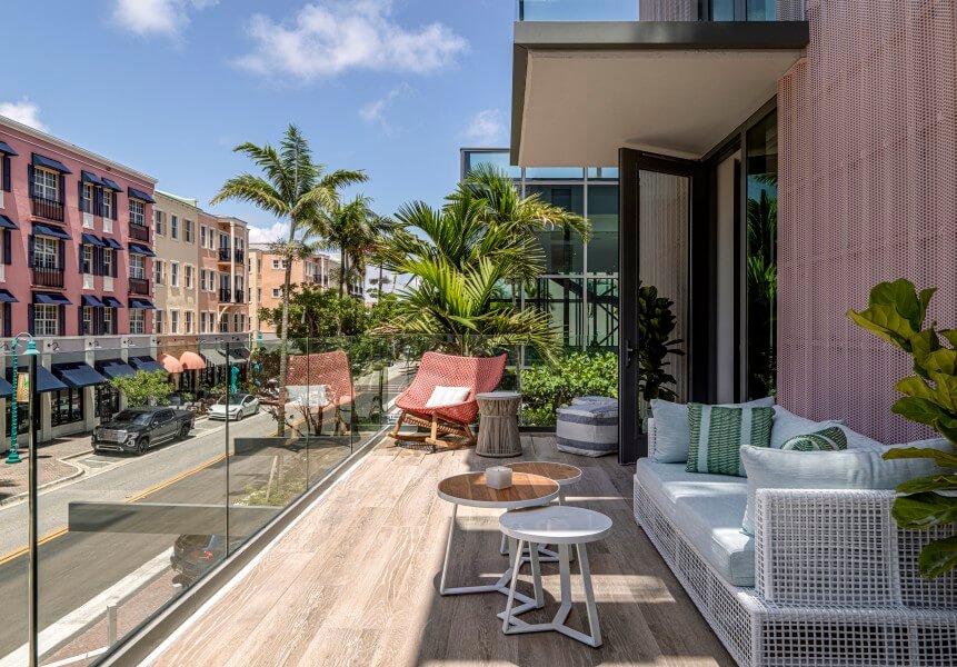 A modern balcony with a white sofa, red chair, and wooden tables overlooks a vibrant street scene with colorful buildings and palm trees