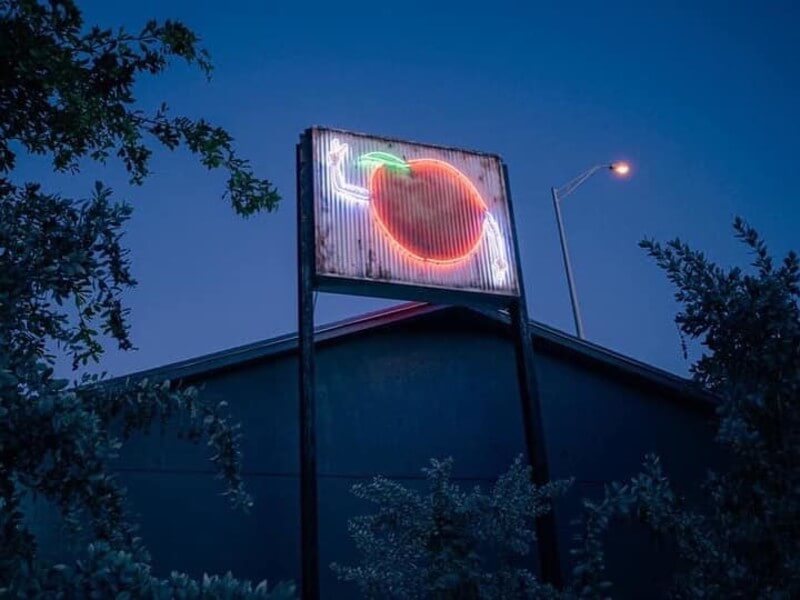 A vintage-style neon sign featuring a glowing red apple with a green stem, mounted on a weathered metal pole against a twilight sky