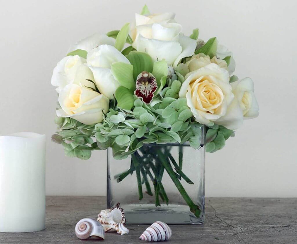 A square glass vase filled with white roses, orchids, and hydrangeas, accompanied by seashells and a white candle