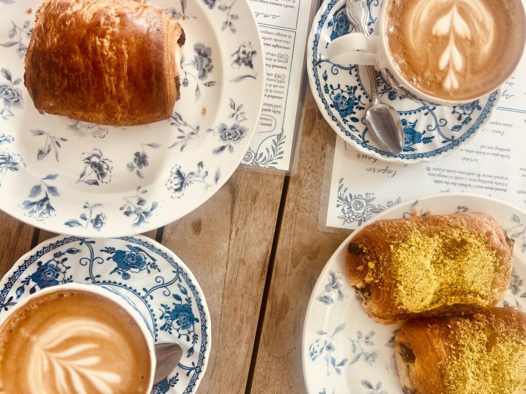 A cozy café scene featuring two golden-brown pastries dusted with powdered sugar, two cups of coffee with intricate latte art, and delicate blue and white floral china on a wooden table