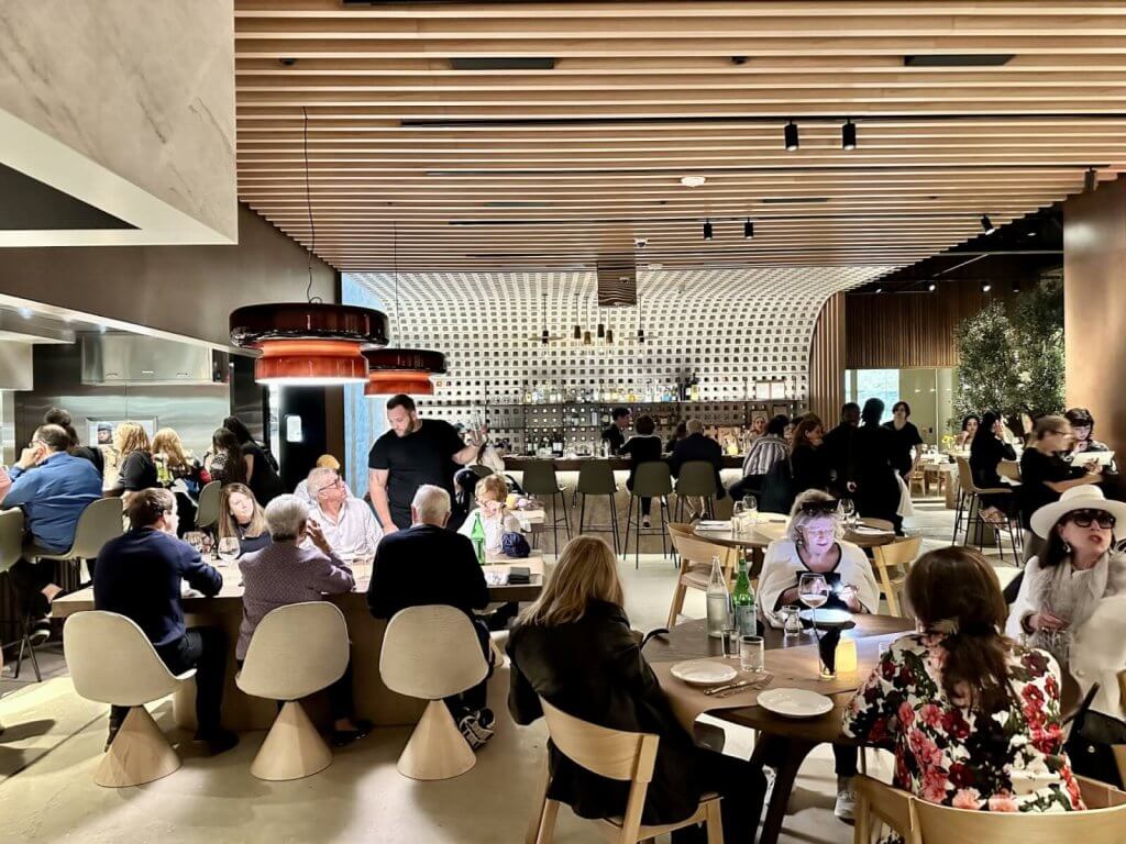 A bustling modern restaurant with a curved wooden ceiling, filled with diners enjoying meals and drinks at tables and the bar