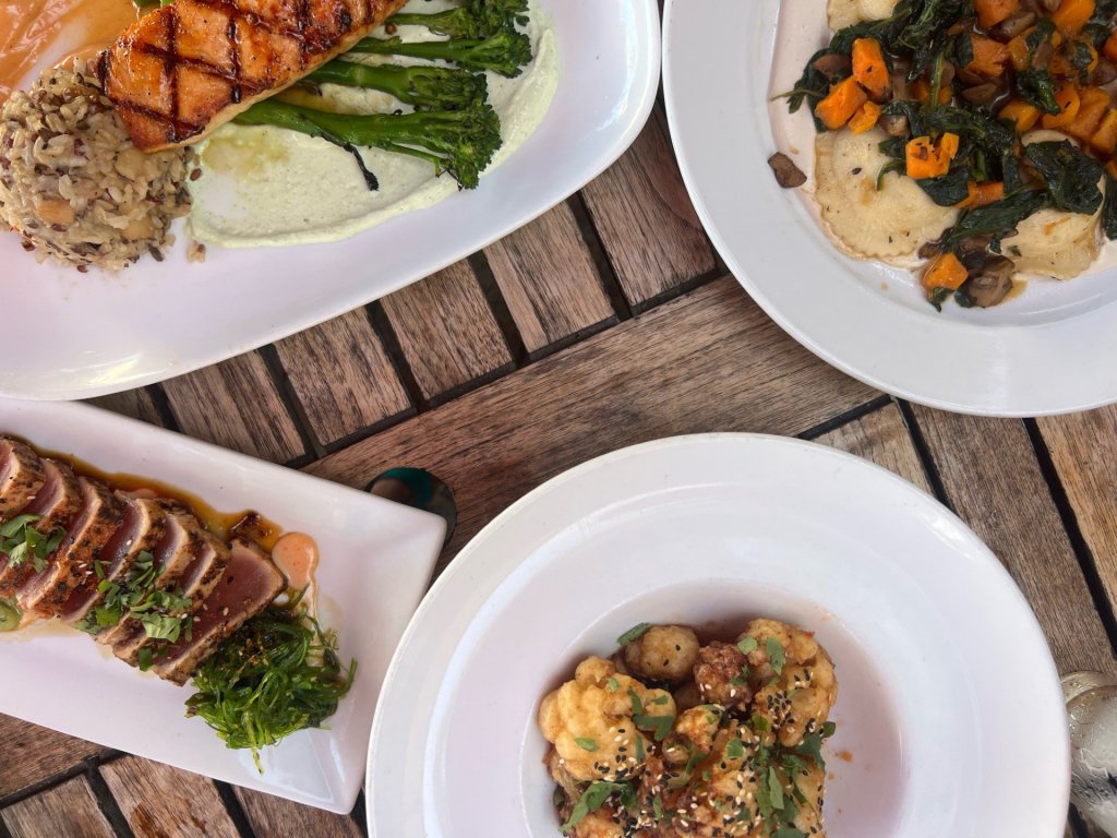 A variety of gourmet dishes including grilled salmon, seared tuna, roasted cauliflower, and a colorful vegetable medley are artfully presented on white plates atop a rustic wooden table