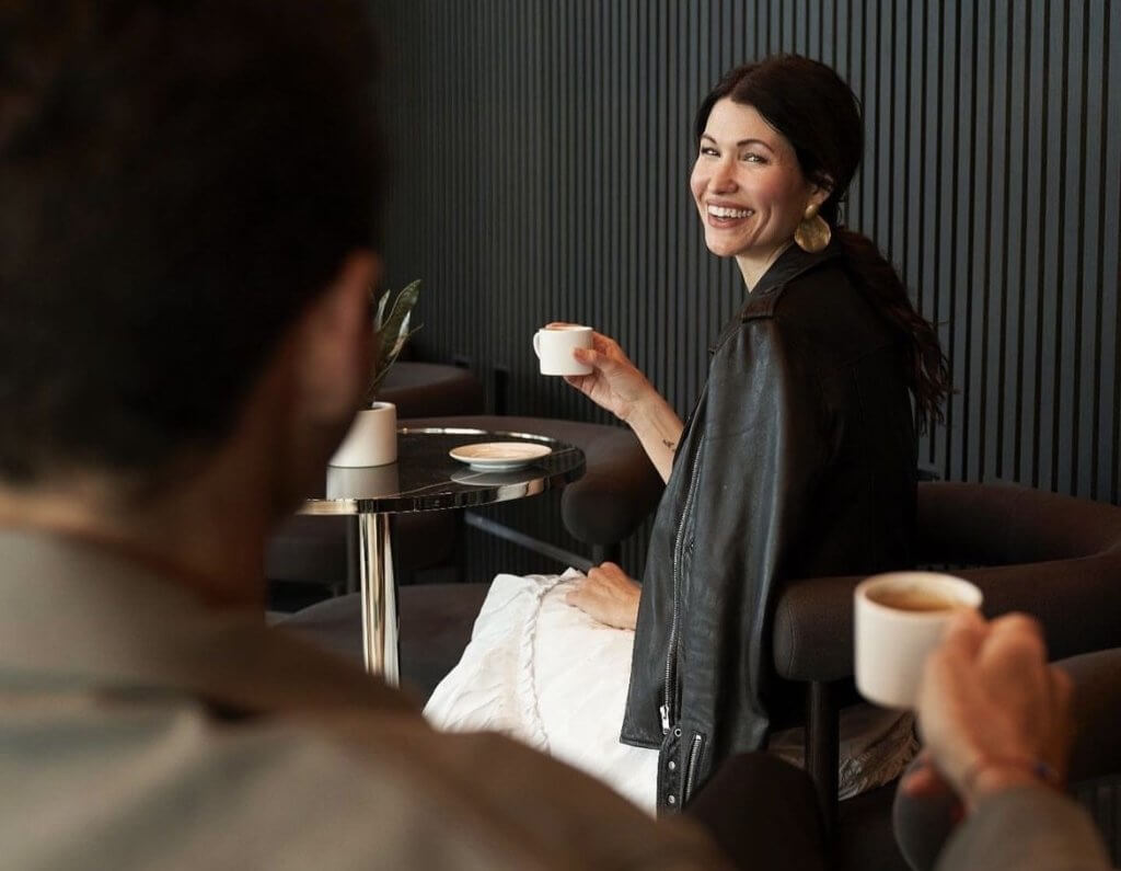 A woman in a black leather jacket smiles brightly while holding a white coffee cup, sitting across from a man at a small round table in a cozy indoor setting