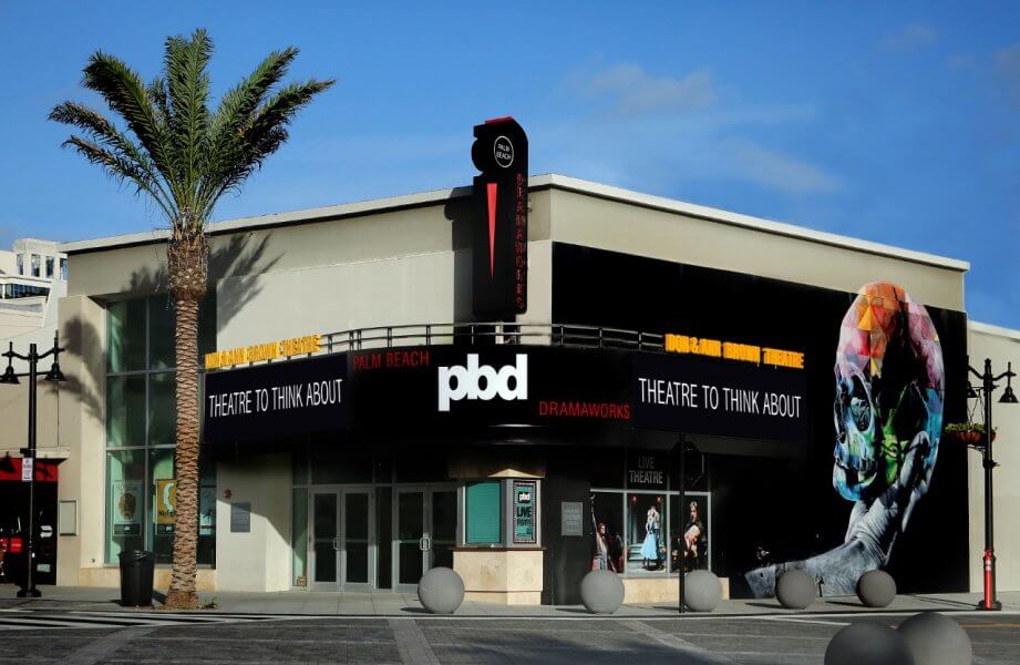 A modern theater building with a large, colorful mural of a skull on its side, featuring the text 'Theatre to Think About' and 'PBD DramaWorks' on its marquee
