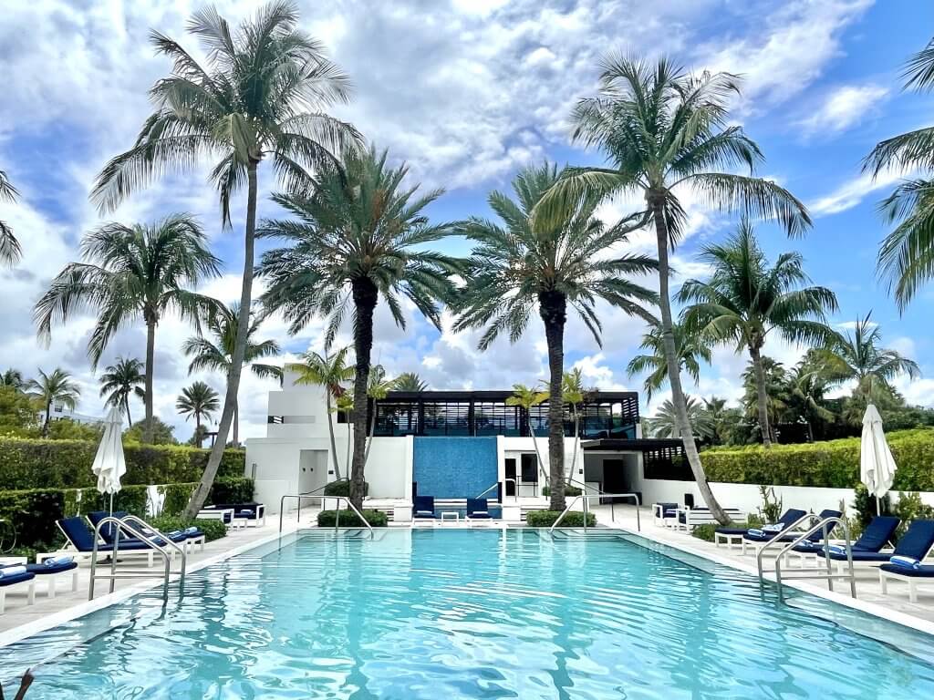 Une oasis tropicale sereine avec une grande piscine accueillante entourée de palmiers luxuriants et de chaises longues blanches modernes.