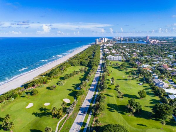 Aerial view of a coastal golf course with lush green fairways, sand traps, and palm trees lining a highway that runs parallel to a pristine beach and the vast blue ocean
